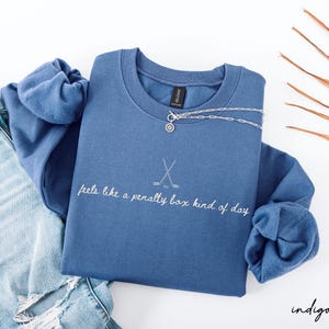 Op de afbeelding: Indigo blauwe sweatshirt met de tekst "feels like a penalty box kind of day" en een hockeystickafbeelding. Een zilveren ketting met een hanger ligt over de sweatshirt. Lichtblauwe jeans zijn zichtbaar.