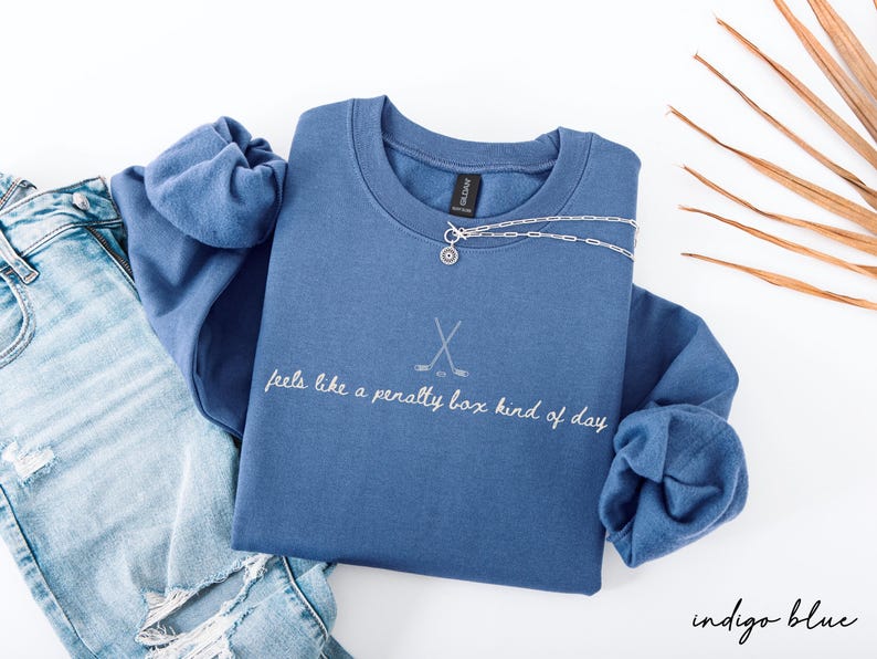 Op de afbeelding: Indigo blauwe sweatshirt met de tekst "feels like a penalty box kind of day" en een hockeystickafbeelding. Een zilveren ketting met een hanger ligt over de sweatshirt. Lichtblauwe jeans zijn zichtbaar.
