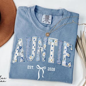 May include: A blue jean Comfort Colors t-shirt with the word "AUNTIE" appliqued in floral and gingham patterns. The shirt includes the text "EST. 2026" and a bow graphic. A gold necklace and a brown hat are also visible.