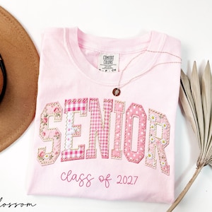 May include: A light pink t-shirt featuring the word SENIOR in large, fabric-appliqued letters with various pink floral, gingham, and polka dot patterns. Below the word, the text class of 2027 is printed in a cursive pink font on the chest.