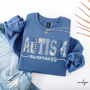 May include: Indigo blue sweatshirt with the word "AUTISM" in patchwork letters and the word "awareness" in script. A silver necklace and a pair of light blue jeans are next to the sweatshirt. A dried palm leaf is in the background.