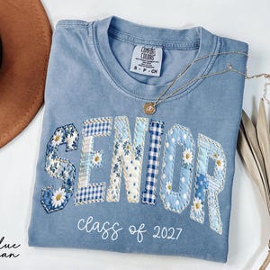May include: A light blue Comfort Colors t-shirt with the word "SENIOR" in patchwork letters, featuring floral and gingham patterns. The text "class of 2027" is below. A gold necklace and a brown hat are also visible.