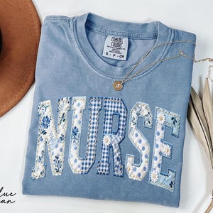 May include: A blue jean Comfort Colors t-shirt with the word "NURSE" appliqued across the front. The letters are a mix of floral, polka dot, and gingham patterns in shades of blue and white. A gold necklace and a brown hat are also visible.