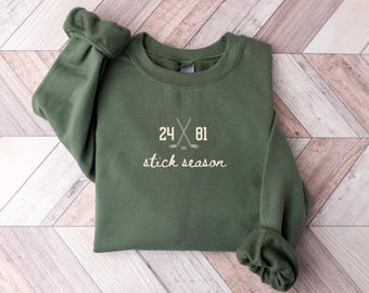 Op de afbeelding: Een olijfgroene sweatshirt met de tekst "24 81 stick season" in het wit. Het ontwerp bevat gekruiste hockey sticks. De sweatshirt is op een licht houtnerf oppervlak gevouwen.