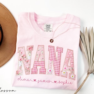 May include: A light pink t-shirt with the word "NANA" in a patchwork design. The letters are a mix of floral, gingham, and polka dot patterns. Below the word are the names "olivia", "james", and "sophie". A gold necklace rests on the shirt.