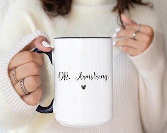 May include: White ceramic mug with a black rim and handle. The mug features the text "DR. Armstrong" in a script font, with a small black heart below the name. The mug is held by a person wearing a white sweater.