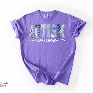 May include: A violet-colored t-shirt with the words "AUTISM awareness" printed on the front. The word "AUTISM" is in a patchwork design with blue and white patterns. The word "awareness" is in a cursive font.