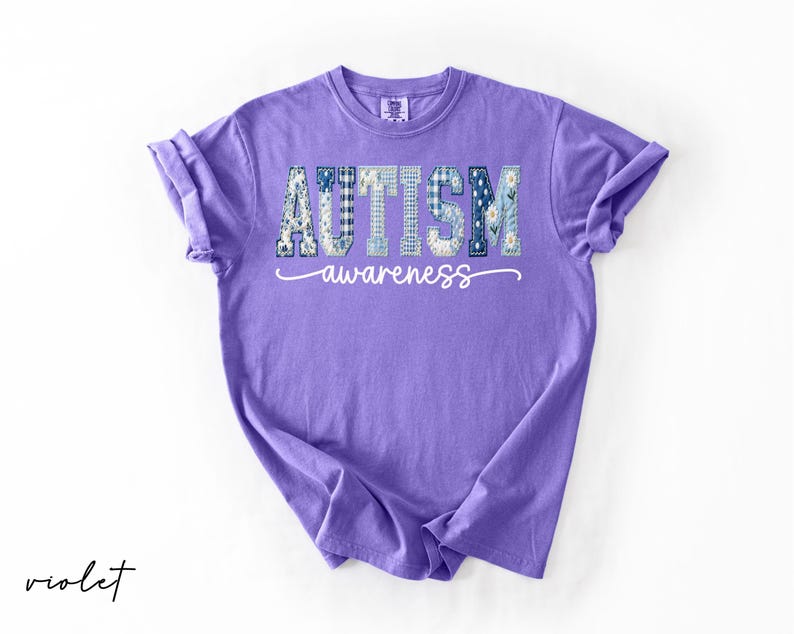May include: A violet-colored t-shirt with the words "AUTISM awareness" printed on the front. The word "AUTISM" is in a patchwork design with blue and white patterns. The word "awareness" is in a cursive font.