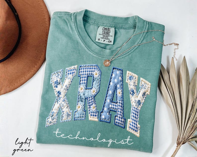 May include: Light green Comfort Colors t-shirt with the word "XRAY" in a patchwork design with blue floral and gingham patterns. The word "technologist" is written below the word "XRAY". A silver necklace with a pendant is on the shirt.