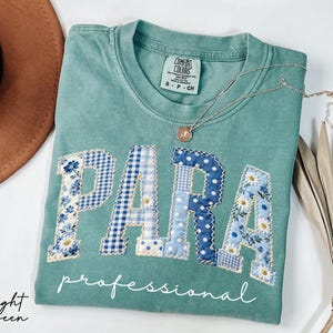 May include: Light green t-shirt with the word "PARA" in patchwork letters, featuring floral and gingham patterns in blue and white. The word "professional" is written below. A silver necklace with a pendant is on the shirt.