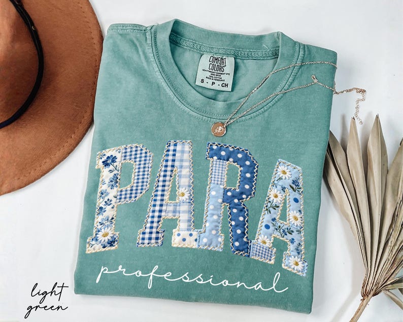 May include: Light green t-shirt with the word "PARA" in patchwork letters, featuring floral and gingham patterns in blue and white. The word "professional" is written below. A silver necklace with a pendant is on the shirt.