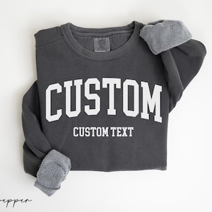 May include: A dark gray sweatshirt with the word "CUSTOM" in large, white, block letters across the chest. Below the word "CUSTOM" is the text "CUSTOM TEXT" in white. The sleeves are folded, revealing a gray, fuzzy lining.