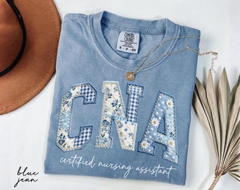 May include: Blue jean colored t-shirt with the letters "CNA" appliqued in floral, gingham, and daisy patterns. The words "certified nursing assistant" are written in script below the letters. A gold necklace and a brown hat are also visible.