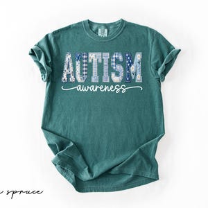 May include: A teal-colored t-shirt with the words "AUTISM awareness" printed across the front. The word "AUTISM" is in a patchwork design with blue and white patterns. The text "awareness" is in a cursive font.