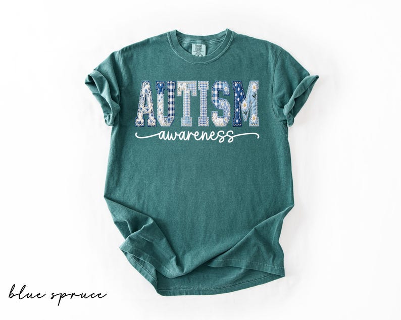 May include: A teal-colored t-shirt with the words "AUTISM awareness" printed across the front. The word "AUTISM" is in a patchwork design with blue and white patterns. The text "awareness" is in a cursive font.