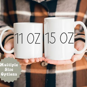 May include: Two white ceramic mugs with black text. The mug on the left says "11 oz" and the mug on the right says "15 oz".