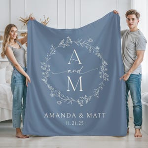 May include: A blue blanket with a white floral wreath design. The wreath surrounds the letters "A" and "M" with the word "and" in cursive. Below the wreath are the names "AMANDA & MATT" and the date "11.21.25".