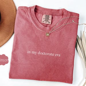 Może przedstawiać: Czerwony t-shirt w kolorze karmazynowym z nadrukiem "in my doctorate era" w kolorze białym na przodzie.