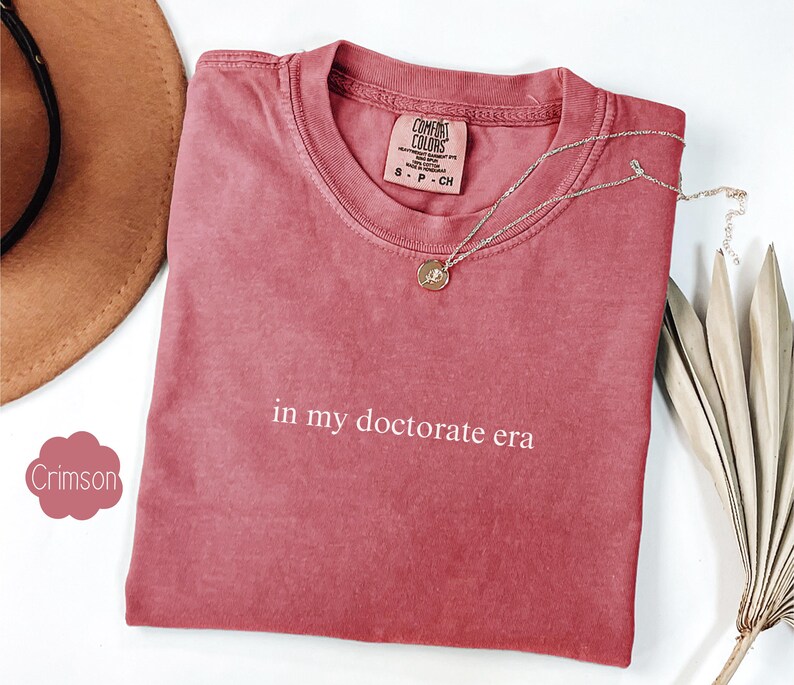 Może przedstawiać: Czerwony t-shirt w kolorze karmazynowym z nadrukiem "in my doctorate era" w kolorze białym na przodzie.