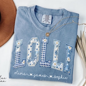 May include: A blue jean Comfort Colors t-shirt with the word "LOLII" in a patchwork design. The letters are a mix of blue gingham, floral patterns, and white daisies. Below the letters are the names "olivia", "james", and "sophie". A gold necklace rests on the shirt.