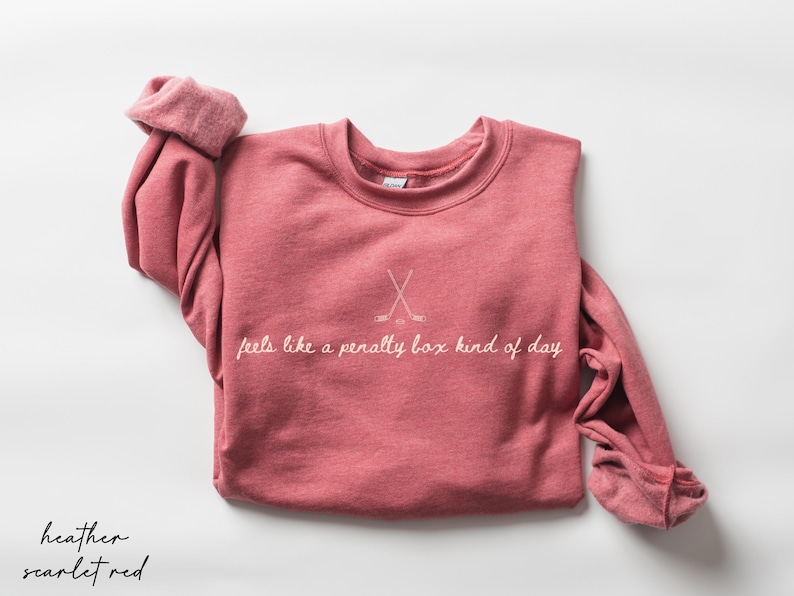 Op de afbeelding: Heather scharlakenrood sweatshirt met de tekst "feels like a penalty box kind of day" en gekruiste hockeysticks. De top met lange mouwen en ronde hals heeft een effen kleur.