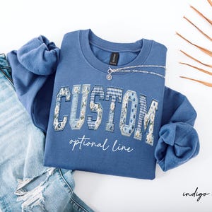 May include: Indigo blue sweatshirt with the word "CUSTOM" in a floral and plaid design. The words "optional line" are below the word "CUSTOM". A silver necklace is on the sweatshirt. Light blue jeans are next to the sweatshirt.