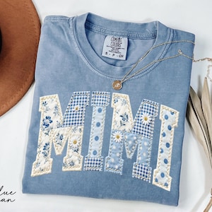 May include: A blue jean Comfort Colors t-shirt with the word "MIMI" appliqued in a patchwork design. The letters feature floral, gingham, and polka dot patterns. A gold necklace with a pendant rests on the shirt.