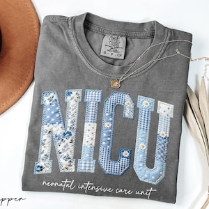 K&ouml;nnte beinhalten: Ein graues Comfort Colors T-Shirt mit den Buchstaben "NICU", die in einem Patchwork-Design aus blauem Blumen-, Tupfen- und Vichy-Stoff appliziert sind. Darunter stehen die Worte "neonatal intensive care unit". Eine goldene Halskette und ein brauner Hut sind ebenfalls zu sehen.