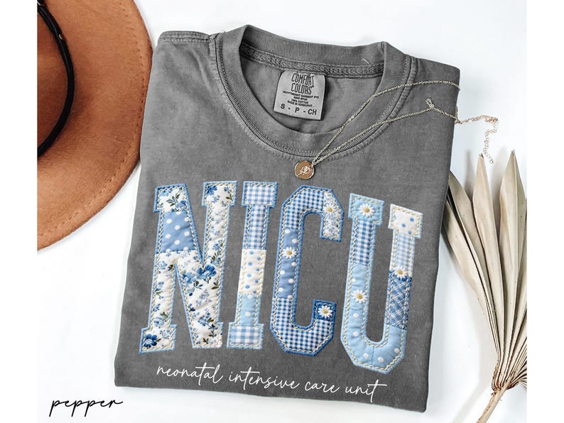 K&ouml;nnte beinhalten: Ein graues Comfort Colors T-Shirt mit den Buchstaben "NICU", die in einem Patchwork-Design aus blauem Blumen-, Tupfen- und Vichy-Stoff appliziert sind. Darunter stehen die Worte "neonatal intensive care unit". Eine goldene Halskette und ein brauner Hut sind ebenfalls zu sehen.