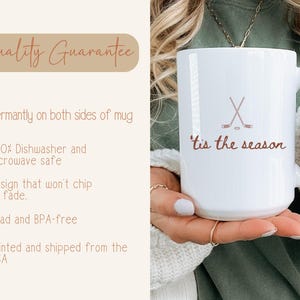 May include: White ceramic mug featuring a hockey stick and puck design, with the words "tis the season" in brown script. The mug has a large handle and is held by a person.