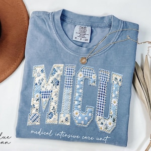 May include: A blue jean Comfort Colors t-shirt with the letters "MICU" appliqued in floral and gingham patterns. The words "medical intensive care unit" are printed below. A gold necklace and a brown hat are also visible.
