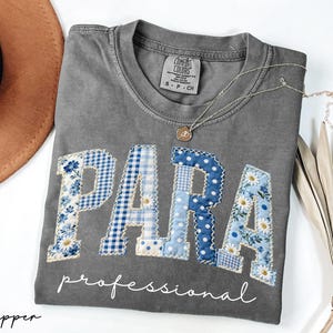 May include: Gray t-shirt with the word "PARA" in blue patchwork letters, with the word "professional" below. The letters have floral and polka dot patterns. A gold necklace and a brown hat are also visible.