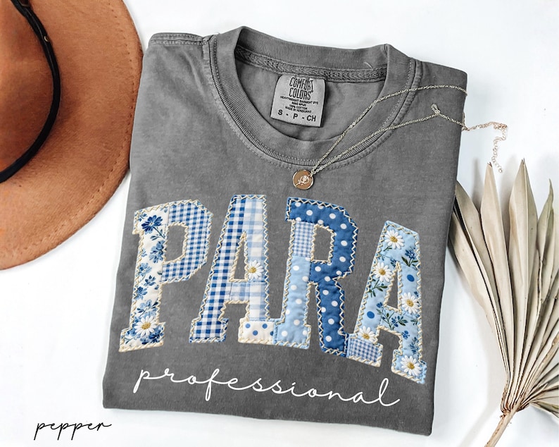 May include: Gray t-shirt with the word "PARA" in blue patchwork letters, with the word "professional" below. The letters have floral and polka dot patterns. A gold necklace and a brown hat are also visible.