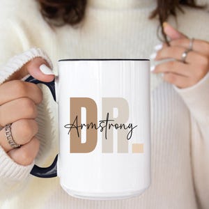 May include: White ceramic mug with a black handle. The mug has the text "DR. Armstrong" in brown and beige lettering.