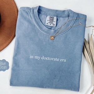 Może przedstawiać: Jasnoniebieska koszulka z nadrukiem "in my doctorate era" w kolorze białym na przodzie. Koszulka jest złożona i leży płasko na białym tle.