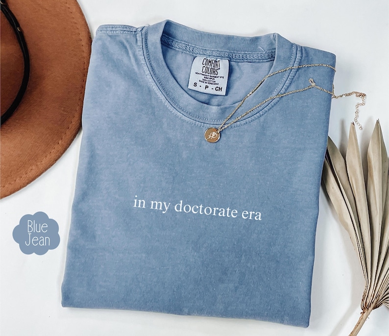 Może przedstawiać: Jasnoniebieska koszulka z nadrukiem "in my doctorate era" w kolorze białym na przodzie. Koszulka jest złożona i leży płasko na białym tle.