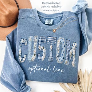 May include: A light blue sweatshirt with the word "CUSTOM" in a patchwork design. The letters are made of floral, gingham, and polka dot patterns. The text "optional line" is written below the word "CUSTOM". A gold necklace is draped over the sweatshirt.