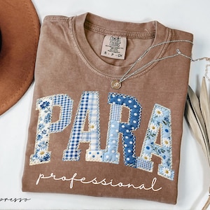 May include: Espresso-colored t-shirt with the word "PARA" in blue and white patchwork letters. The word "professional" is written below in cursive. A silver necklace and a brown hat are also in the image.
