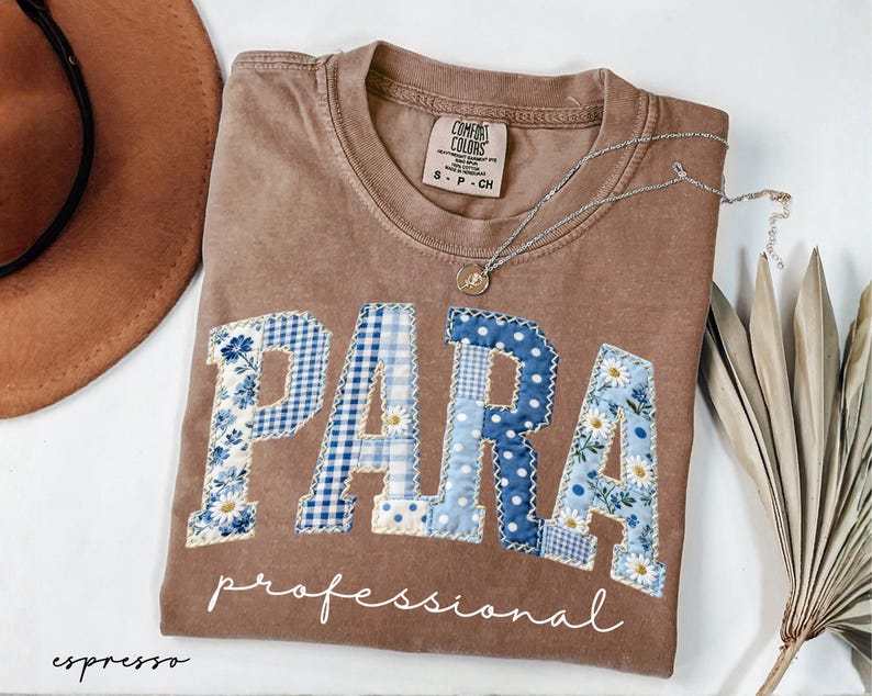 May include: Espresso-colored t-shirt with the word "PARA" in blue and white patchwork letters. The word "professional" is written below in cursive. A silver necklace and a brown hat are also in the image.