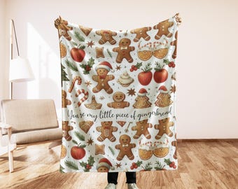 May include: A white blanket with a repeating pattern of gingerbread men, apples, orange slices, and holiday greenery. The blanket has the text "You're my little piece of gingerbread" in a script font.
