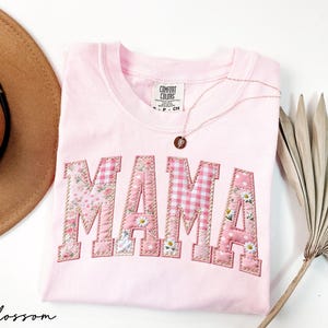 May include: A light pink t-shirt with the word "MAMA" appliqued in a patchwork design. The letters feature floral, gingham, and polka dot patterns. A gold necklace with a pendant rests on the shirt. A brown hat and dried palm leaf are in the background.