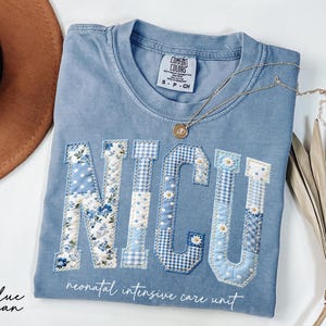 K&ouml;nnte beinhalten: Hellblaues Jeans-T-Shirt mit den Buchstaben "NICU", die mit verschiedenen blauen Blumen- und Vichy-Mustern appliziert sind. Die Worte "neonatal intensive care unit" sind darunter aufgedruckt. Eine goldene Halskette mit Anh&auml;nger liegt auf dem Shirt.