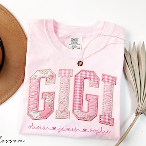 May include: A light pink Comfort Colors t-shirt with the name "GIGI" appliqued in pink floral and gingham fabric. The names "olivia", "james", and "sophie" are embroidered below. A gold necklace and a brown hat are also visible.
