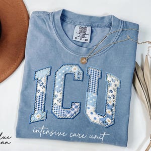 May include: A blue jean Comfort Colors t-shirt with the letters "ICU" appliqued in patchwork fabric with floral and gingham patterns. The words "intensive care unit" are printed below the letters. A gold necklace rests on the shirt.