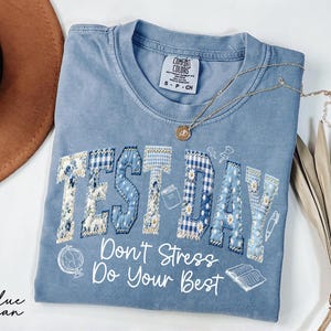 May include: A blue jean Comfort Colors t-shirt with the words "TEST DAY" in a patchwork design. The shirt also features the text "Don't Stress Do Your Best" and small illustrations of a globe, book, and pencil.