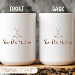 May include: Two white ceramic mugs with black handles and rims. Each mug features a crossed hockey stick design and the phrase "'tis the season" in brown script. The mugs are displayed on a wooden surface.