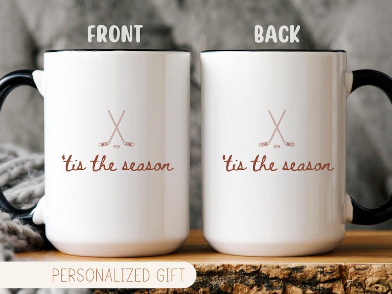 May include: Two white ceramic mugs with black handles and rims. Each mug features a crossed hockey stick design and the phrase "'tis the season" in brown script. The mugs are displayed on a wooden surface.