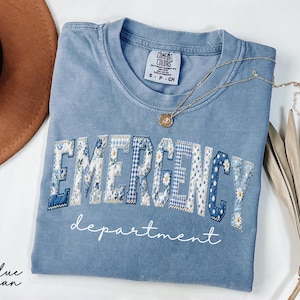 May include: A blue jean Comfort Colors t-shirt with the words "EMERGENCY department" in a patchwork design. The letters are a mix of floral, gingham, and polka dot patterns. A gold necklace and a brown hat are also visible.