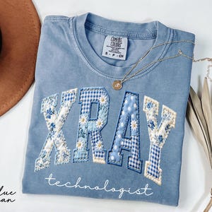 Light blue Comfort Colors® t-shirt featuring "XRAY" in floral patchwork letters with technologist script underneath, designed for radiology professionals and medical staff.
