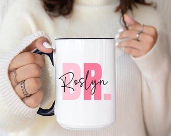 May include: White ceramic mug with a black handle. The mug has a pink and white graphic that says "DR. Roslyn".